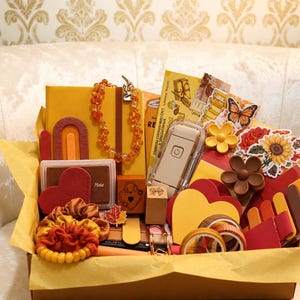 May include: A gift box filled with stationery and accessories in shades of yellow, orange, and red. Items include a yellow notebook, a beaded necklace, a heart-shaped stamp pad, a small electronic device, floral stickers, hair scrunchies, and washi tape.