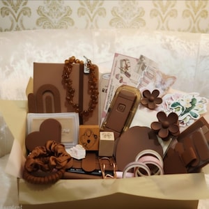 May include: A collection of brown-toned stationery and accessories including a notebook, a beaded necklace, a heart-shaped stamp, a hair claw, a scrunchie, washi tape, and various stickers arranged in a box with light-colored tissue paper.