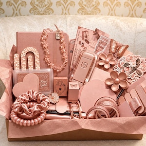 May include: A gift box filled with rose gold items including a notebook, a beaded necklace, a hair scrunchie, a spiral hair tie, a small portable fan, butterfly and flower clips, heart-shaped cards, washi tape, and various stationery accessories.