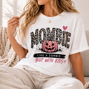 May include: A white t-shirt featuring the word MOMBIE in a brown and black leopard print pattern. Below the text is a pink jack-o'-lantern pumpkin graphic. The text reads LIKE A ZOMBIE, BUT WITH KIDS, accompanied by a small pink heart icon.