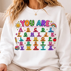May include: A white crewneck sweatshirt featuring a colourful graphic of the alphabet from A to Z. Each letter is paired with a positive adjective, such as Accepting for A and Zealous for Z. The design includes icons like a sun, rainbow, pencil, and books.