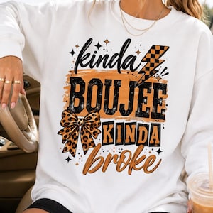 May include: A white long-sleeve sweatshirt featuring a graphic print with the text "kinda BOUJEE KINDA broke" in black and orange. The design includes a leopard print bow, a checkered lightning bolt, and small star accents in a distressed style.