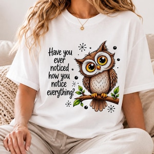 May include: A white short-sleeved t-shirt featuring a brown owl perched on a branch with large yellow eyes. The text reads, Have you ever noticed how you notice everything? The design includes small black star-like accents and green leaves.