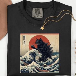 May include: A black t-shirt featuring a graphic print of a black cat resembling a monster rising from a stylized ocean wave, with a large red sun, Mount Fuji, and Japanese text in a vertical box on the left side of the rectangular illustration.