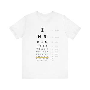 May include: A white short-sleeved t-shirt featuring a black eye chart graphic on the front. The chart displays rows of letters and numbers, including the text I, NB, RIG, HTE, STDAYI, NBLACK, ESTNIGH, TNOEVILS, HALLESCAP, EMYSIGHT, and visual acuity fractions.