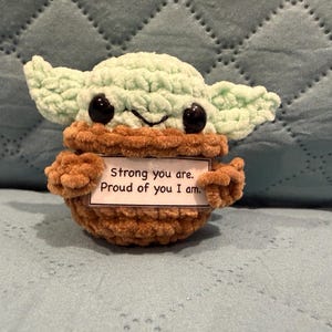 Op de afbeelding: Een klein gehaakt wezen met een lichtgroene huid, grote zwarte ogen en puntige oren. Het draagt een bruin gehaakt gewaad en houdt een wit rechthoekig bordje vast met de tekst, Strong you are. Proud of you I am. Het figuur heeft een zachte textuur.