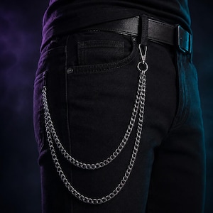 May include: A silver-toned double-layered metal wallet chain attached to a black leather belt and black denim jeans. The chain features two distinct loops of interlocking metal links that hang from a single lobster clasp hook at the belt loop.