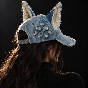 Peut inclure: Une casquette de baseball en jean bleu délavé dotée de deux oreilles pointues et dressées aux bords effilochés. La casquette présente un motif découpé décoratif sur le côté et une sangle réglable avec une boucle en métal à l'arrière.