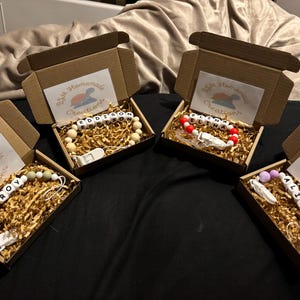 May include: Four open brown cardboard boxes containing personalised dummy clips with white letter beads spelling EDDISON, ELINA, ROY, and ALICIA. Each box includes gold paper filler and a white card with the text B&M Homemade Creations.