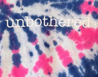 May include: A short-sleeve V-neck t-shirt featuring a blue and pink spiral tie-dye pattern. The word unbothered is printed in white lowercase letters across the center of the chest. The shirt has a Gildan brand label at the inner neckline.