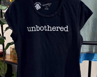 May include: A black short-sleeved t-shirt featuring the word unbothered printed in white across the chest. The shirt has a crew neckline and a tag on the inside collar that reads Stray Cotton and Universal Thread with a small circular logo.