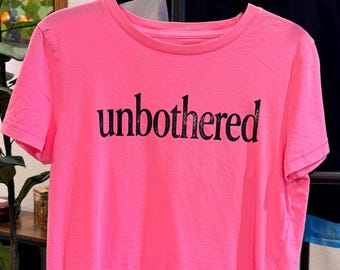 May include: A bright neon pink short-sleeved t-shirt featuring the word unbothered printed in black, serif-style lettering across the center of the chest. The garment has a classic crew neckline and is displayed on a black hanger.