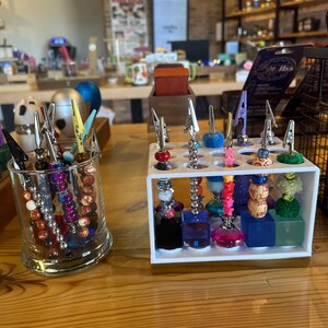 May include: A collection of metal alligator clips attached to decorative beaded rods, organized in a clear glass cylinder and a white rectangular stand with square colored bases, featuring various bead shapes, colors, and textures.