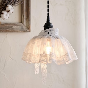 May include: A pendant light fixture featuring a black cord and a small black bird figure attached to the top. The light is covered by a white lace lampshade with a ruffled edge and two hanging lace ribbons, emitting a soft warm glow from within.