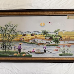 Puede incluir: Un bordado en seda enmarcado de forma rectangular que representa un paisaje rural con un río, bambú y figuras con sombreros cónicos. La escena incluye un pequeño bote, búfalos de agua y un sol dorado. El marco es marrón oscuro con borde dorado.