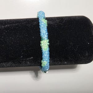 May include: A cylindrical beaded bracelet featuring a repeating pattern of light blue and lime green glass seed beads. The beads are arranged in a dense, tubular weave, creating a textured surface with a consistent diameter throughout the length.