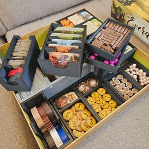 May include: A black board game organiser insert for the Everdell game box, featuring multiple compartments filled with game components including stacks of cards, brown wooden logs, yellow circular tokens, pink berries, and white pearl beads.