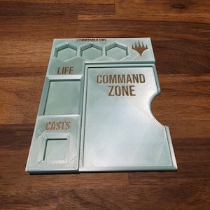 以下が含まれることがあります： Magic: The Gathering用のライトミントグリーンの3Dプリント製ゲームボード。COMMAND ZONE、LIFE、CASTSとラベル付けされたセクション、COMMANDER DMG用の3つの六角形の溝、右上の金色のロゴが特徴です。