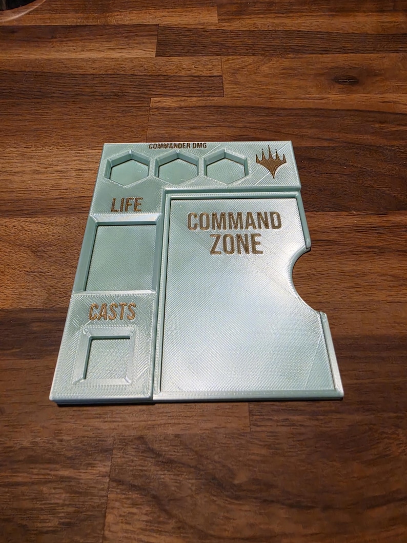 以下が含まれることがあります： Magic: The Gathering用のライトミントグリーンの3Dプリント製ゲームボード。COMMAND ZONE、LIFE、CASTSとラベル付けされたセクション、COMMANDER DMG用の3つの六角形の溝、右上の金色のロゴが特徴です。
