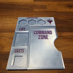 May include: A light blue 3D-printed game board accessory featuring recessed slots and purple text. The board includes sections labelled COMMAND ZONE, LIFE, and CASTS, with a top row for COMMAND DMG and a Magic: The Gathering symbol.