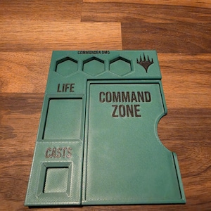 以下が含まれることがあります： COMMAND ZONEとラベル付けされた大きな長方形のエリア、LIFE用のセクション、CASTS用のセクション、シンボルの上の3つの六角形の溝を備えたティール色のプラスチック製ゲームボードアクセサリーで、上部にCOMMANDER DMGというテキストが印刷されています。