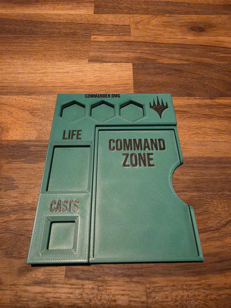 以下が含まれることがあります： COMMAND ZONEとラベル付けされた大きな長方形のエリア、LIFE用のセクション、CASTS用のセクション、シンボルの上の3つの六角形の溝を備えたティール色のプラスチック製ゲームボードアクセサリーで、上部にCOMMANDER DMGというテキストが印刷されています。