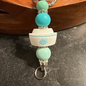 May include: A keychain featuring a white nurse cap charm with a light blue cross, accented by mint green and turquoise round beads, silver-toned rhinestone spacers, and a metal lobster clasp and ring at the top and bottom for attachment.
