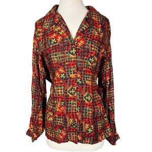 May include: A long-sleeved button-down shirt featuring a repeating pattern of red, yellow, and black heraldic shields with fleur-de-lis and lion motifs set against a dark, textured diamond-shaped background with a classic collar and buttoned cuffs.