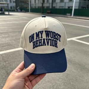 May include: A baseball cap with a cream-colored crown and a navy blue brim. The front of the crown features the text "ON MY WORST BEHAVIOR" embroidered in navy blue, bold, arched lettering. The cap has a structured five-panel design.