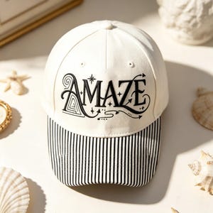 May include: A cream-colored baseball cap featuring the word AMAZE embroidered in black on the front panel. The cap has a structured crown with a button on top and a curved brim decorated with a vertical black and white pinstripe pattern.