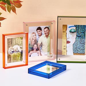 May include: A collection of four rectangular acrylic photo frames in orange, pink, green, and blue. The orange frame holds a puppy photo, the pink frame displays a family portrait, the green frame shows an abstract face illustration, and the blue frame lies flat.