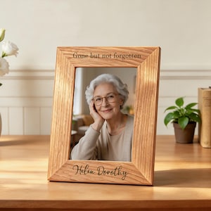 May include: A rectangular light brown wooden picture frame with a visible grain texture. The top border is engraved with the text "Gone but not forgotten" and the bottom border features the engraved name "Helen Dorothy" in script font.