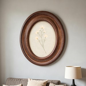 May include: An oval-shaped wall frame with a dark brown wood grain finish. The frame contains a light cream-coloured mat with a centred botanical illustration of a delicate, thin-stemmed plant featuring small, pale white flowers and green leaves.