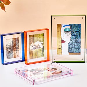 Peut inclure: Un ensemble de quatre cadres photo rectangulaires en acrylique bleu, orange, vert et rose. Trois cadres sont disposés verticalement avec de l'art abstrait, un chiot et un visage stylisé, tandis qu'un cadre rose est posé horizontalement avec une photo.