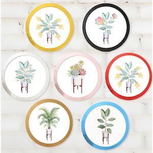 May include: A collection of seven circular wall art pieces featuring watercolour illustrations of various potted plants. Each piece is framed in a different coloured border, including gold, black, white, pink, red, and blue, displaying botanical art.