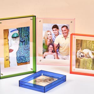May include: A collection of four rectangular acrylic photo frames in vibrant colours including green, pink, orange, and blue. Each frame features a clear centre panel held by small metal corner screws, displaying various photographs and art prints.