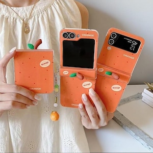 Könnte beinhalten: Drei orangefarbene Handyhüllen für ein klappbares Smartphone, jeweils mit Punktmuster, einem kleinen grünen Blattakzent und einem weißen ovalen Etikett mit dem Aufdruck "Orange". Eine Hülle ist mit einem hängenden Anhänger mit Perlen versehen.