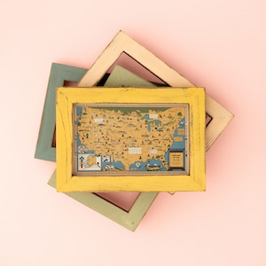 Puede incluir: Un marco de madera amarillo con un mapa vintage de los Estados Unidos en su interior. El mapa est&aacute; rodeado por un borde azul claro y presenta varias ilustraciones de monumentos y ciudades. El marco est&aacute; superpuesto a otros dos marcos, uno verde claro y otro marr&oacute;n claro.
