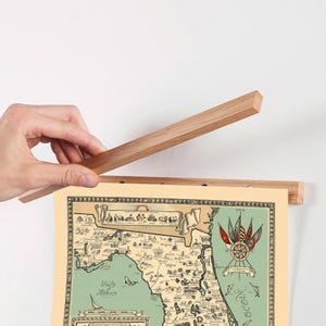 Pu&ograve; includere: Una cornice magnetica in legno chiaro per poster che sostiene una stampa di una mappa della Florida in stile vintage. La mappa presenta uno sfondo color crema, una massa terrestre verde e illustrazioni decorative di bandiere, navi e testo.