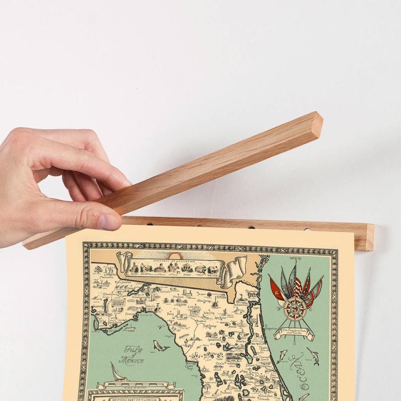 Pu&ograve; includere: Una cornice magnetica in legno chiaro per poster che sostiene una stampa di una mappa della Florida in stile vintage. La mappa presenta uno sfondo color crema, una massa terrestre verde e illustrazioni decorative di bandiere, navi e testo.