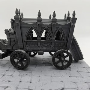 May include: A dark gray miniature hearse carriage featuring gothic arched windows, pointed finials, and skull motifs along the roofline. The carriage body is adorned with bat carvings and rests on four spoked wheels, with a coffin attached at the rear.