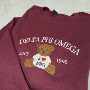 May include: A maroon crewneck sweatshirt featuring white embroidered text that reads DELTA PHI OMEGA, EST 1998, and a brown teddy bear wearing a white t-shirt with a red heart and the Greek letters Delta Phi Omega embroidered on the front.