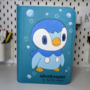 May include: A rectangular blue binder featuring a cartoon penguin character with a yellow beak and feet, surrounded by white bubbles and star-like accents. The text MintKeeper by The Arty Woopah! is printed in black at the bottom right corner.