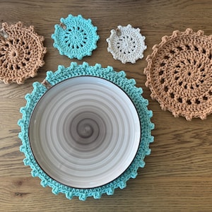 Op de afbeelding: Vier gehaakte onderzetters in bruin en turquoise, met een grotere turquoise gehaakte placemat en een wit bord in het midden.