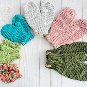May include: A set of six crocheted mittens in various colors, including teal, green, gray, pink, and a multi-colored pattern. The mittens are arranged in a semi-circle on a white wooden surface.