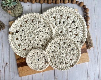 Könnte beinhalten: Ein Set aus vier cremefarbenen, handgehäkelten Tischsets und einem passenden Untersetzer. Die Tischsets haben ein kreisförmiges, florales Design und sind aus dickem, strukturiertem Garn gefertigt. Das Set wird auf einem Holzschneidebrett präsentiert.