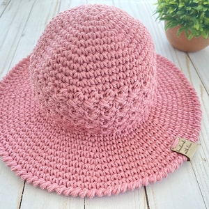 May include: A dusty rose crochet sun hat with a wide brim. The hat is handmade, as indicated by a small tag that reads "handmade with love." The hat's texture is visible, with a detailed stitch pattern. The hat is designed for sun protection.