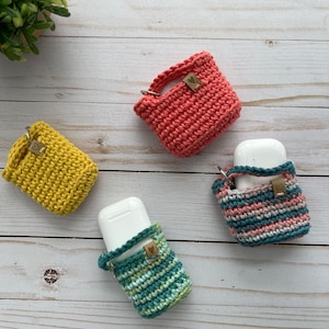 Op de afbeelding: Vier gehaakte AirPod-hoesjes in verschillende kleuren: koraal, geel, teal en roze en witte strepen. Elk hoesje heeft een klein hartvormig label.
