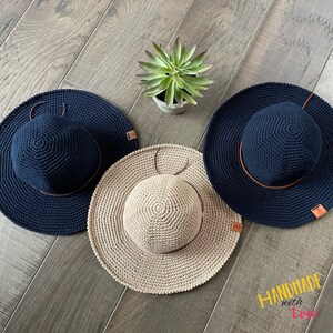 May include: Three crocheted sun hats, one beige and two navy blue, with brown leather bands. The hats are arranged on a wooden surface with a small potted succulent plant in the center.