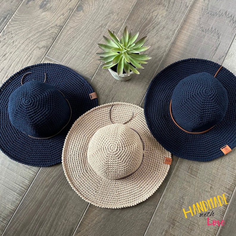 May include: Three crocheted sun hats, one beige and two navy blue, with brown leather bands. The hats are arranged on a wooden surface with a small potted succulent plant in the center.